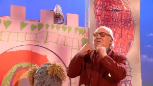Een foto van meneer Aart in bruin pak met een bruine bril op en het achterhoofd van pop Tommy, waarvan de oren uit een soort zilverkleurige helm op zijn hoofd steken. Ieniemienie staat op een soort getekende kasteelmuur met een witte punthoed op.
