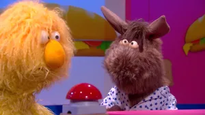 Een foto van de pop Ian van Balken en de pop Opper de Pop, die achter een rode drukknop staan. Ian heeft een bruine vacht, met omhoogstekende oren zoals een Ezel. Ian draagt een witte blouse met blauwe sterren erop. Opper's vacht is geel.