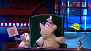 Een foto van de pop van Nico de Neushoorn op een groene stoel achter een bureau met een opengeslagen boek erop. Naast pop Nico is de pop Stella Schaap te zien. Stella heeft een bord vast met daarop een getekende bruine boterham.