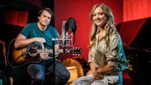 Een foto van Sterre die op een kruk zit, voor een zwarte piano en achter een microfoon. Ze lacht naar de camera. Linksachter zit Bernard met een akoestische gitaar. op de achtergrond is een stukje van een trommel van een drumstel te zien achter een lamp.