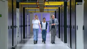 Een foto van Tess, Aditya en een man die in een gang van een gebouw lopen. In de gang staan links en rechts acht paarse metalen kasten met twee deuren, die dichtzitten. Tussen iedere kast zit  steeds loopruimte om ernaast te kunnen lopen. 