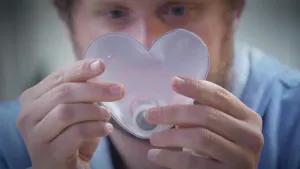 Een foto van Diederik, die in zijn handen een hart van doorzichtig plastic vastheeft en ernaar kijkt. In het hart zit een rond, wit dingetje met een grijze zijkant. Het is een handwarmer, om koude handen mee te verwarmen.