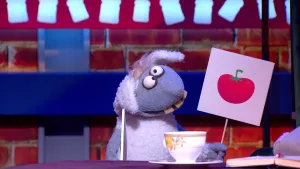 Een foto van pop Stella Schaap. De pop heeft een bord met een getekende rode tomaat erop vast en kijkt naar het bord. De pop heeft een witte vacht op haar rug en oren, een grijze vacht bij kop en poten, ronde ogen en vooruitstekende, gele tanden.