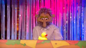 Een foto van pop Jantje van Leiden in een witte blouse en met een lange neus, die naar een plastic eend met oranjerode snavel kijkt op de boterhamband. De band zelf is niet te zien, maar de muur eronder met getekende stukken kaas, tomaat en sla erop wel.