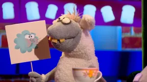 Een foto van pop Stella Schaap met een bord met een getekende blauwe wolk die lucht blaast erop. De pop kijkt naar het bord. De pop heeft een witte vacht op rug en oren, een grijze vacht bij kop en poten, ronde ogen en vooruitstekende, gele tanden.