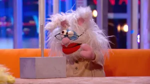 Een foto van pop Elvira Einstein op een bruine bank achter een tafel. Op de tafel staat een soort vierkant betonblok, waar een metalen staaf uit omhoog steekt. Elvira heeft pluizig wit haar, konijnenoren, schele ogen, een bril en grote, rode lippen.