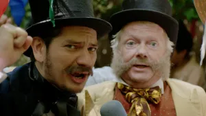 Een foto van twee mannen met een zwarte, hoge hoed. Eén draagt een donkerblauw pak, kijkt in de camera, heeft zijn vuist omhoog en een microfoon vast. De ander draagt een lichtbruin pak, een donkerbruin vest met witte stippen en een goudbruine strik.