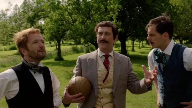 Een foto van drie mannen op een grasveld met bomen erachter. De middelste man houdt een bruine voetbal met één hand tegen zich aan. Zijn andere hand houdt hij met gekromde vingers voor zijn lichaam. De mannen dragen een jasje en stropdas.
