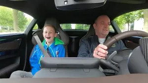 Een foto van Stéphan achter het stuur van een personenauto. Thijs zit er lachend naast op de bijrijdersstoel. Ze rijden door een straat of over een weg met bomen aan de kant.