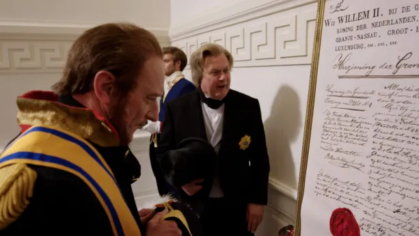 Een foto van twee verklede mannen bij een groot document in een goudkleurige lijst. Op het document staat "Willem II" en de tekst "Herziening der Grondwet". De man verkleed als Koning Willem II draagt een goudkleurig lint met blauwe strepen over zijn pak.