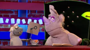 Een foto van pop Stella Schaap met vooruitstekende, gele tanden. De pop heeft een bord vast met een getekende sleutel erop en kijkt ernaar. De pop staat naast de groene stoel van pop Nico de Neushoorn die met een bril op ook naar de tekening kijkt. 