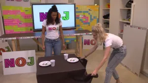 Een foto van Sosha en Janouk bij een vierkante tafel met twee borden en twee bekers erop, die op een bruin tafelkleed staan. Janouk pakt twee punten van het tafelkleed vast op de foto.