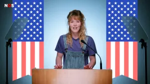 Een foto van een presentator achter een lichtbruine spreektafel met een microfoon erop. Op de achtergrond zijn twee getekende, Amerikaanse vlaggen te zien, deels met rode en witte strepen en deels met witte sterren op een blauwe achtergrond.