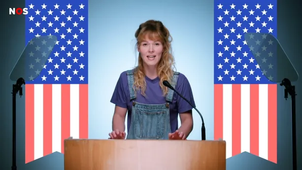 Een foto van een presentator achter een lichtbruine spreektafel met een microfoon erop. Op de achtergrond zijn twee getekende, Amerikaanse vlaggen te zien, deels met rode en witte strepen en deels met witte sterren op een blauwe achtergrond.