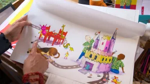 Een foto van de handen van Mylo Freeman,  die één van haar tekeningen vastheeft en ernaar wijst. Op de tekening spelen twee jongetjes bij een groot kasteel met in hun handen een ridder en er liggen en staan knuffels, poppetjes, een hond en een kat op.