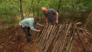 Een foto van twee mensen op een grond van aarde met op de achtergrond bomen. Ze zijn bezig om een rij van houten paaltjes van boomtakken tegen elkaar aan te zetten. De paaltjes staan aan twee kanten schuin tegen een lange, houten paal aan.