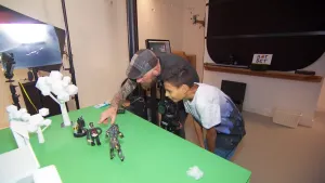 Een foto van Damien en Jorn die bij een groene tafel staan. Jorn wijst naar een poppetje met lange armen op de tafel. Ernaast is een ander poppetje op de rug te zien. Op de tafel staan ook witte boomachtige vormen en links een witte vorm van een gebouw.