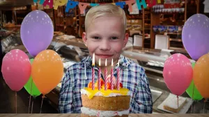 Een foto van een jongen achter een plaatje van een verjaardagstaart met vijf brandende kaarsjes erop. Links en rechts zijn vier ballonnen te zien. Achteraan hangt een slinger met "Happy Birthday" en ligt een taart in een koelvitrine in een taartenwinkel.