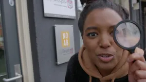 Een foto van presentator Sosha die buiten voor een deur van een bedrijf staat. Ze staat recht voor de camera en heeft een vergrootglas recht voor haar linkeroog. 