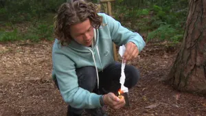 Een foto van presentator Pascal die op de grond zit in het bos. Hij houdt een stuk wc-papier vast met zijn linkerhand en steekt die aan met een aansteker in die hij in zijn rechterhand vast heeft.