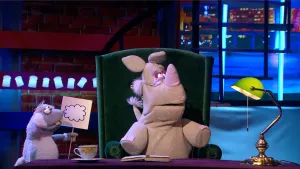 Een foto van Nico de Neushoorn die achter een tafel in een stoel zit. Voor hem ligt een open boek op tafel en ernaast een kop en schotel. Naast hem staat er een witte pop met een bord zijn handen waar een wolk op getekend staat.