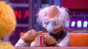 Een foto van pop professor Bolleboos. Hij heeft een beker voor zich op tafel vast en heeft de vingers van zijn andere hand tegen de bovenrand van de beker liggen. De beker is wit van binnen en heeft strepen van verschillende kleuren van buiten.