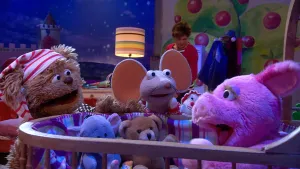 Een foto van de pop Purk, die in een houten wieg met knuffels erin zit. Tommie, met een wit en rood gestreepte slaapmuts op, en Ieniemienie staan naast de wieg. Purk heeft haar mond open. Achteraan staat Lot naast een lamp op een tafel.