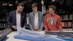 Een foto van modeontwerper Janice, Teun en Tim die achter een witte tafel staan. Janice pakt een rol vast met een lichtblauwe stof erop. Ook ligt een andere rol die nog ingepakt is met plakband eromheen en een rechthoekig deel van een kledingstuk.