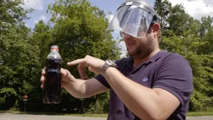 Een foto van Diederik die buiten staat met een plastic kap op zijn hoofd en deels voor zijn gezicht. Hij heeft een doorzichtig plastic flesje vast met bruine frisdrank, cola, erin. Een laagje butagas dat Diederik er eerder opgegoten heeft, drijft erop.