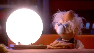 Een foto van pop professor Bolleboos met grijs haar, een bril en een rimpelig gezicht. Hij heeft zijn handen gevouwen op de tafel liggen en kijkt naar een lichtgevende, bolvormige lamp. De lamp zit aan een rechthoekig ding met drie groene knopjes erop.