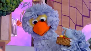 Een foto van pop Pino met een rieten mandje. Uit het mandje steekt een groen blad en een gele paardenbloem. Pop Pino heeft een lichtblauw verenpak, een oranje snavel en twee ronde ogen met gele veren erachter.