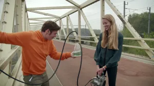 Een foto van twee mensen op een fiets- en loopbrug. Een man houdt een slang achter zich vast die aan een ding met vocht erin zit in zijn andere hand. Het ding zit met een andere slang aan een apparaat met twee ronde metertjes dat een vrouw vastheeft.