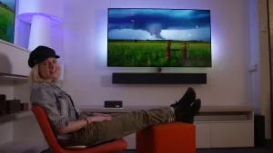 Een foto van presentator Janouk die in de camera kijkt terwijl ze achterover in een stoel zit. Op de achtergrond is er een televisie te zien die aanstaat.