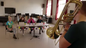 Een foto van presentator Janouk die naast een jury zit die iemand beoordelen die op de voorgrond staat en trompet speelt.