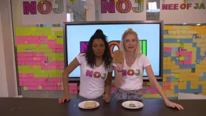 Een foto van presentatoren Sosha en Janouk die in de studio achter een tafel staan. Janouk laat een boterham met pindakaas erop zien en er staat een leeg bord voor haar. Voor Sosha staat er een bord met een snee brood erop.