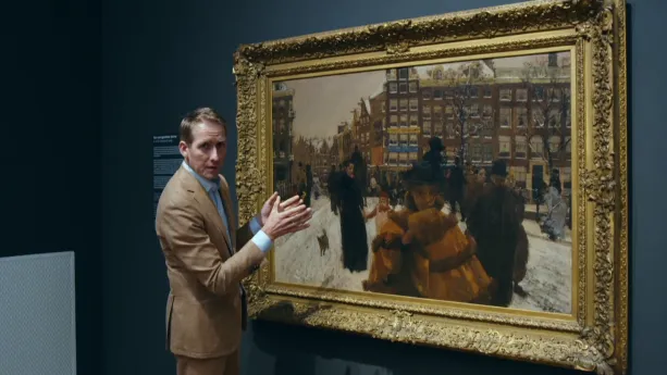 Een foto van een man die in een museum voor een schilderij staat en daarover wat aan het uitleggen is.