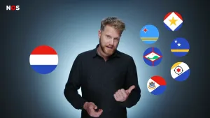 Een foto van een presentator die voor de camera staat. Links van hem is er een Nederlandse vlag. De presentator wijst met zijn duim naar de vlaggen rechts van hem, het zijn de vlaggen van Aruba, Bonaire, Curaçao, Saba, Sint Eustatius en Sint Maarten.