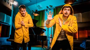 Een foto van presentatoren Buddy en meester Willem die in de studio staan. Ze hebben beide gele regenjassen aan en Buddy kijkt angstig naar boven.