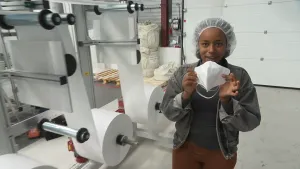 Een foto van een presentator die een mondkapje in haar handen heeft en die laat zien voor de camera. Op de achtergrond staat er een machine met twee grote rollen met een soort papier waarvan de vellen buiten beeld gaan..