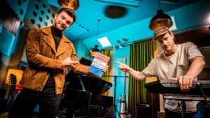 Een foto van presentatoren Buddy en meester Willem in de studio met allemaal muziek instrumenten om hun heen. Buddy houdt een grondwet boek vast en beide wijzen daarnaar.