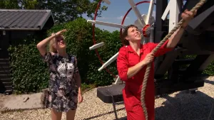 Een foto van Janouk die omhoogkijkt in de lucht. Ernaast staat molenaar Marloes in een rood werkpak. Ze trekt aan een dik, bruin touw dat rechtsboven aan een niet-zichtbaar deel van de molen vastzit. Erachter is een wit met rood wiel te zien.