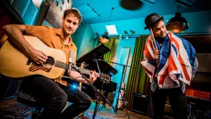 Een foto van meester Willem op een stoel met zijn hand op de snaren van een akoestische gitaar. Buddy heeft de vlag van het Engels sprekende Groot-Brittannië om zijn nek hangen, draagt een zwarte bolhoed en krabt op zijn hoofd met een stok, een drumstok.