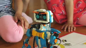 Een foto van een robot van lego op een papier met een niet duidelijk zichtbare tekening erop. De robot heeft lichtblauwe, zwarte, witte en oranje plastic stukjes, met ronde, lichtblauwe ogen. Op de buik zitten drie gele stukjes in de vorm van een strik.