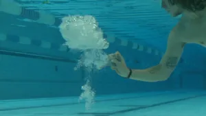 Een foto van Pascal in een zwembad onder water. Hij heeft iets in zijn hand wat lijkt op een blauwe schaar en ernaast zijn allemaal bubbels in het water te zien. Hij heeft net een gele ballon kapotgeprikt met de schaar in de aflevering.