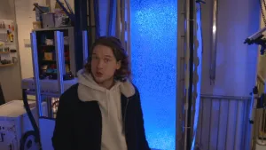 Een foto van Pascal die naast een rechthoekige, hoge bak staat met metalen buizen aan beide zichtbare zijkanten. In de bak zijn allemaal bubbels te zien in het water. De bak wordt verlicht door blauw licht.