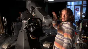 Een foto van Pascal die bij een grote camera staat. De camera zit met gekleurde kabels aan een grote, metalen draaiarm en de grote, ronde zwarte lens wijst iets naar beneden in de richting van de camera die gebruikt is om deze foto te nemen. 