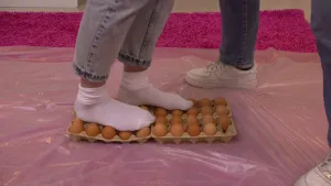 Een foto van de benen van Janouk en Sosha. Janouk loopt met witte sokken over vier kartonnen dozen met bruine eieren heen. Eén voet staat op één rij eieren, de andere voet staat voor de helft op twee rijen. Eronder ligt plastic over het paarse vloerkleed.