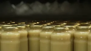 Een foto waarop glazen potten met een goudkleurige deksel van de zijkant te zien zijn. In de potten zit appelmoes. De potten staan in een fabriek en er zit nog geen sticker of label op de potten.