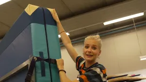 Een foto van Janouk die een metalen rek met een dikke mat die gebruikt wordt voor gymnastiek erop vastheeft in een grote ruimte. De mat zit door een zwarte band vast op het rek. De mat is groen van kleur met geelachtige hoeken.