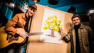 Een foto van meester Willem met een acoustische gitaar en Buddy die met een stift naar een letter "d" op een tekening op een houten bord wijst. Buddy heeft muzieknoten in rondjes op een groengebladerde boom getekend. Tussen de rondjes lopen lijntjes. 